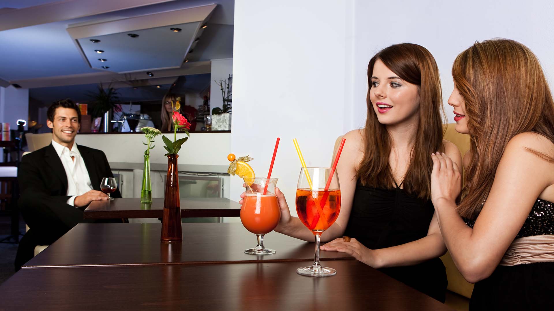 Brunette man in black suit facing two brunette women on the table opposite from him who are enjoying two cocktails.